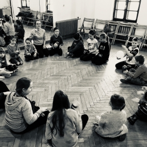Zdjęcie czarno białe, sala z lustrami.Grupa dzieci siedzi na podłodze tworząc okrąg.