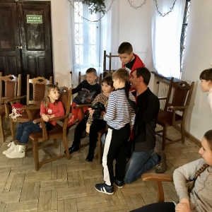 Sala z brązowymi krzesłami, na których siedzi grupa dzieci.Wśród nich mężczyzna z aparatem fotograficznym.