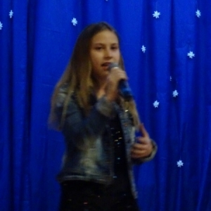 Na tle jasnoniebieskiej ściany stoi dziewczyna , trzyma mikrofon, śpiewa.
