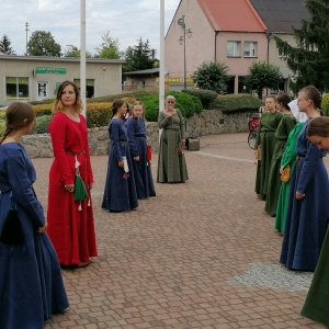 Zespół kobiet w średniowiecznych strojach stoi w dwóch rzędach, w oddali kobieta trzymająca mikrofon.